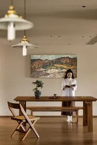 Woman standing near a wooden dining table with a painting, two chairs, and a potted plant