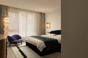 A well-lit bedroom with a large bed, two chairs, and black and white color scheme, featuring curtains on the windows