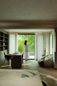 A woman stands near a window in a well-lit room with a wooden floor, two chairs, a couch, and a teddy bear on the floor