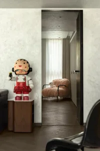 A small living area with a doll on a chair, a teddy bear on the floor, and a couch in the background, creating a cozy and inviting atmosphere
