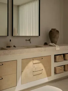 A modern bathroom with a white theme and wooden elements, featuring a sink, mirror, and storage cabinet, with two bottles on the countertop and bright inviting lighting