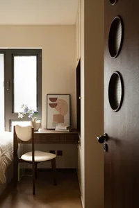 A calm bedroom with a white bed, two chairs, a desk, and a book, with a window nearby