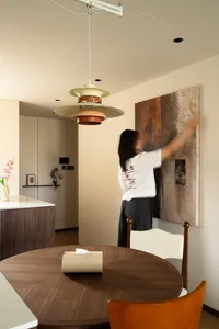 A woman cleans a painting in a kitchen with a dining table, chairs, vases, and a potted plant