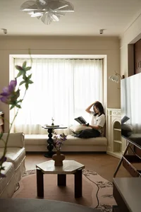 A woman sits on a couch in a cozy living area with a dining table, potted plant, and vases, enjoying a book and the indoor atmosphere