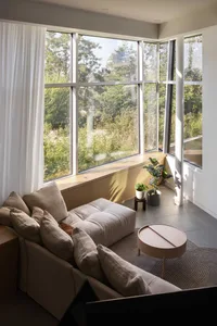 A bright and inviting living area with a couch, chair, and large windows, featuring two potted plants and a comfortable furniture arrangement