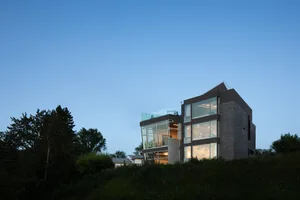 A modern house on a hillside with a combination of glass walls and brick facade, featuring multiple levels, windows, and a rooftop terrace