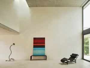 空曠極簡客廳，白色牆面、水泥地板、兩把椅子、一張長凳，以及牆上畫作
