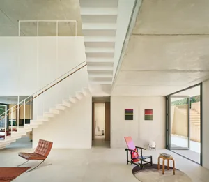 Open modern living area with a white staircase, two chairs, a couch, and natural light from windows, set against a neutral color palette and concrete walls