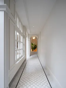 A well-lit hallway with white walls, a tile floor, and two large windows on each side, featuring a potted plant near one of the windows