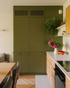 A kitchen with green cabinets, wooden floors, and a dining table, surrounded by potted plants and decorative vases