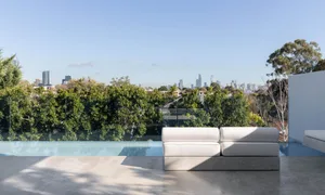 A modern outdoor living area with a white couch on a concrete slab in front of a glass wall with a city view, surrounded by trees