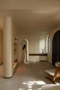 A cozy living area with a woman standing in a hallway between two rooms, featuring two chairs and a potted plant, creating a warm and inviting atmosphere