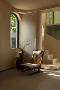 A wooden chair sits on a white carpet in front of a window, surrounded by two potted plants, creating a calm and inviting atmosphere