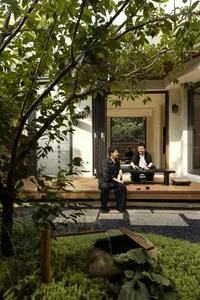 A casual outdoor patio scene with two men seated at a dining table under a tree, surrounded by wooden benches and a relaxed atmosphere