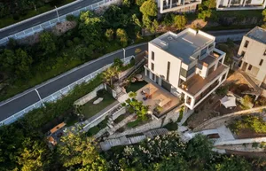 A modern house with a white and gray facade, situated on a hillside surrounded by trees, blending into the environment