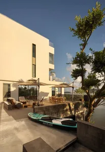 Outdoor living area with a white building, patio, chairs, dining table, umbrellas, and a boat on a lake
