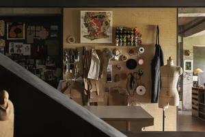 A cluttered workspace filled with clothes, hats, scissors, and tools, with a suitcase and dresses scattered throughout, and numerous hats on various surfaces