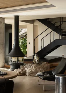Cozy living area with a black fireplace, a staircase, a rock wall, and a potted plant, featuring two chairs and a book on the floor