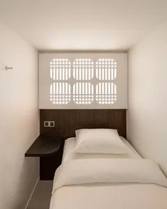 A modern bedroom with a white comforter and brown headboard, a desk, and a chair, well-lit by natural light through a window