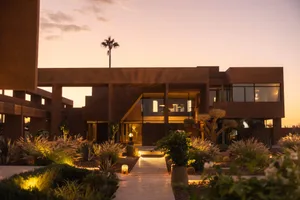 A modern desert-style house with a mix of stone and wood materials, surrounded by palm trees and well-lit with natural light from the windows