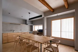 A modern kitchen and dining area with a wooden table and chairs, a refrigerator, oven, and large dining table, surrounded by natural light and sunlight