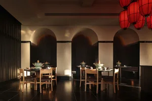 A modern restaurant with a dining area featuring a long table and chairs, and a kitchen with an oven and sink, set against a predominantly white background with dim lighting