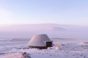 A serene snow-covered landscape with a small wooden hut or shelter in the distance, surrounded by snow and bathed in soft, warm sunlight