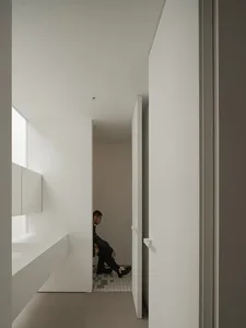 A man sits on a toilet in a small, white bathroom with a tile floor, wearing a black jacket