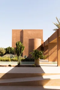 A modern villa with a tan exterior, surrounded by a garden with cacti and a large patio with a pool, set against a bright and natural light background