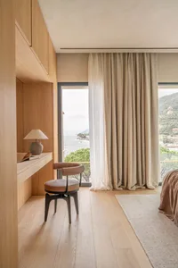 A modern bedroom with a bed, chair, and desk, featuring a large window with natural light and a view of the outdoors