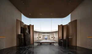 A modern office space with a curved glass wall, a large atrium, and a blue sculpture, surrounded by a circular seating area, illuminated by natural light