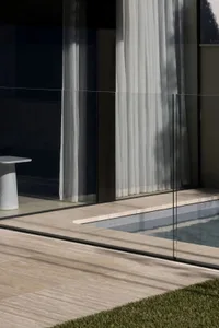 A modern pool area with a glass door and wall, surrounded by a patio, illuminated by natural light, creating a bright and inviting atmosphere