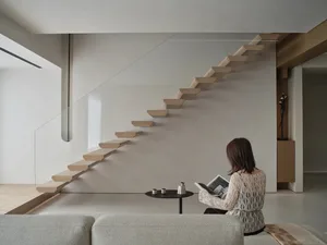 A woman sits on a couch in a well-lit living room with a staircase and a table with a cup and book