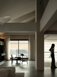 Woman walking towards a dining table in a modern living room with a couch, lamp, and white and grey colors, dim lighting creating a cozy atmosphere