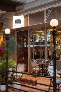 A dimly lit restaurant with a mix of wood and metal materials, featuring tables and chairs, and patrons dining and socializing