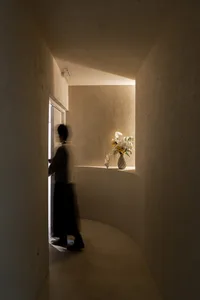 A calm hallway with a woman walking down it, featuring concrete walls, a vase with flowers on a shelf, and a serene atmosphere
