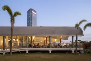 A modern restaurant with wooden dining tables and chairs, set against a backdrop of a tall skyscraper, with soft and warm lighting creating a sophisticated atmosphere