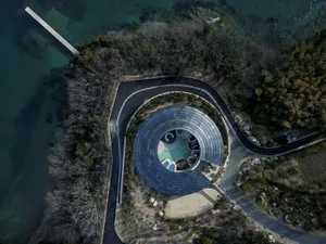 Photograph of a modern house with a circular pool house surrounded by trees and situated near a body of water