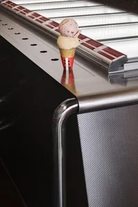 A close-up photo of a chocolate ice cream cone on a silver surface, with a refrigerator in the background