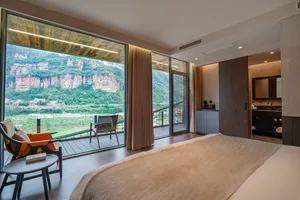 Cozy modern bedroom with a bed, chair, and couch, featuring a large window with a view of the outdoors, wood flooring and furniture, and a white comforter on the bed