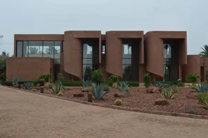 A modern building with a unique design, surrounded by a lush courtyard with cacti and palm trees, featuring red and brown colors and a curved structure