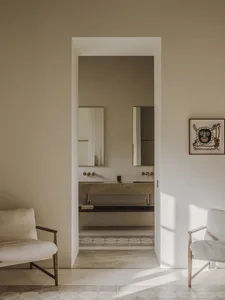 A clean and organized residential bathroom with a white tile floor, sink, mirror, and chair, lit by natural sunlight