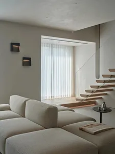 A modern living room with a couch and chair, featuring a glass staircase and large window, with natural light and a clean, minimalist appearance