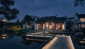 A serene and inviting Chinese garden setting with a large ornate building, a courtyard, and a pond, surrounded by stone architecture and lush greenery
