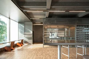 Modern open-plan kitchen and living space with a wooden floor, large window, glass table, and metal bar
