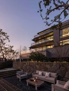A modern building with a stone wall and patio area, featuring a glass wall, two couches, and a chair, creating a comfortable outdoor seating area with natural light and a warm atmosphere