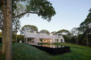 A modern house with a wooden exterior, a large pool surrounded by grass and trees, set against a warm sunset backdrop