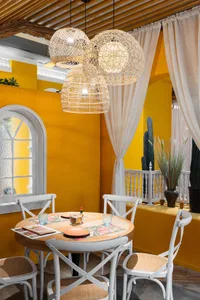 Dining room with a yellow wall, a table set with plates, cups, and bottles, surrounded by white chairs, a tablecloth, and a potted plant