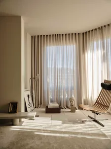 A modern living room with a large window, a couch, chair, dining table, and bookshelf, all in a predominantly white space with natural light