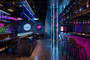 A modern bar with a black counter and pink floor, surrounded by chairs and a dining table in a dimly lit environment